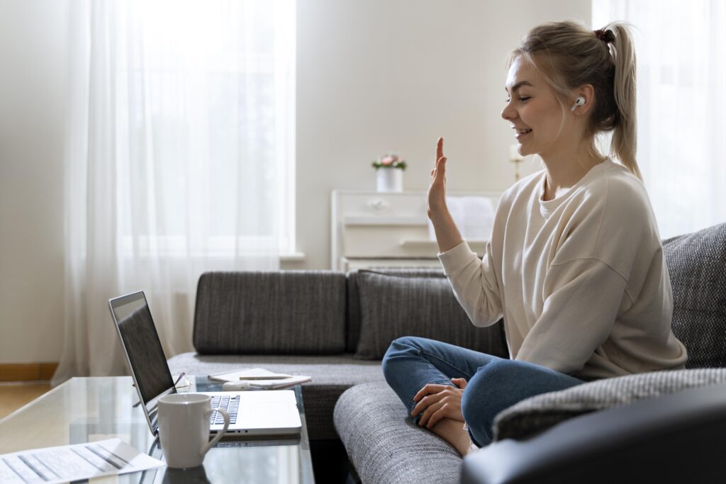 Young woman sitting on a couch at home, smiling and waving during an online therapy session on her laptop, representing mental health counselling.